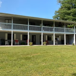 Two-story building with a covered porch on both levels, featuring rocking chairs and potted plants. The building is surrounded by a well-maintained grassy lawn and trees under a clear blue sky.