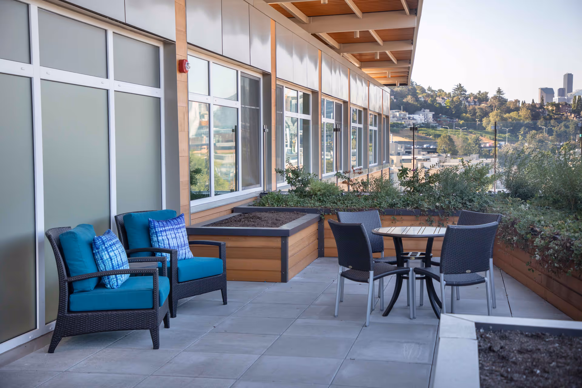 Outdoor patio area with two cushioned wicker chairs with blue pillows and a round table surrounded by four black chairs. The patio is adjacent to a building with large windows and planter boxes filled with greenery. In the background, there is a view of a hillside with houses and some city buildings.
