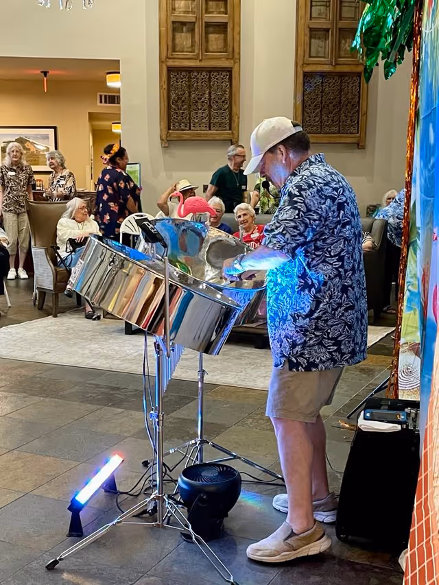 A musician plays a steel drum while senior residents sit and watch in a communal lounge.