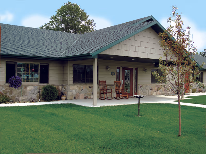 Front entrance of a one-story assisted living building with a covered porch, rocking chairs, stone siding, and a manicured lawn.