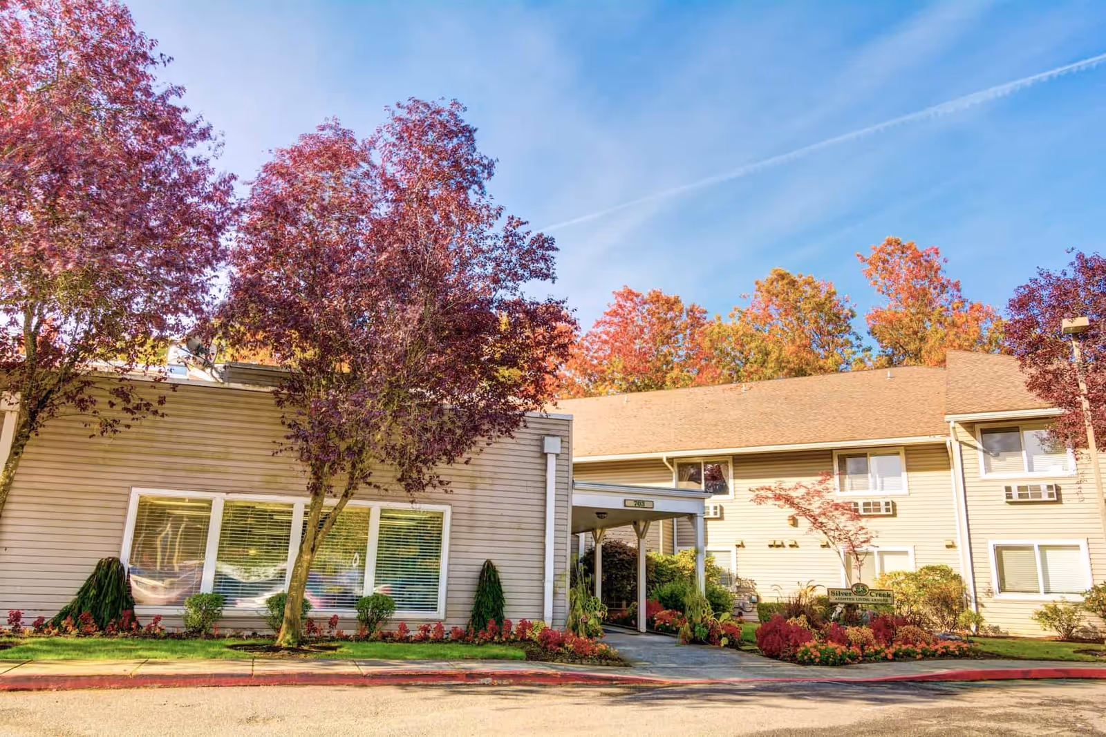 Exterior view of Silver Creek senior living facility with beige siding, large windows, and a covered entrance. The building is surrounded by well-maintained landscaping including red and green bushes and trees with red and orange autumn leaves under a clear blue sky.