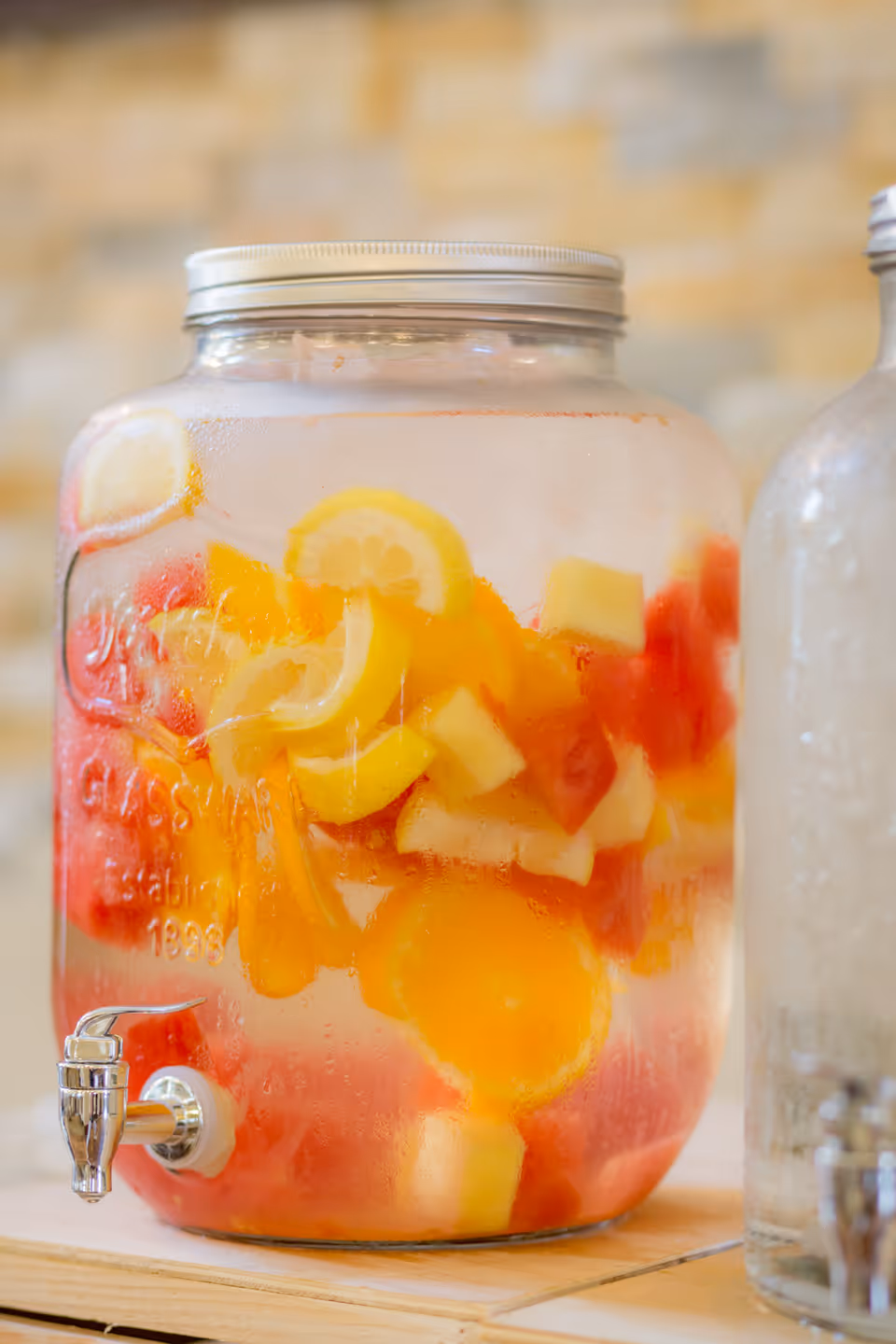 A large glass beverage dispenser filled with water and various sliced fruits including lemons, oranges, and watermelon, placed on a wooden surface with a blurred brick wall background.