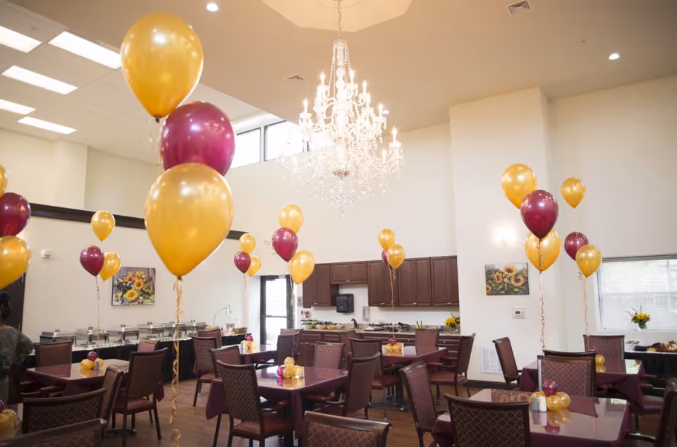 A decorated dining room with a crystal chandelier, tables and maroon and gold balloons throughout.