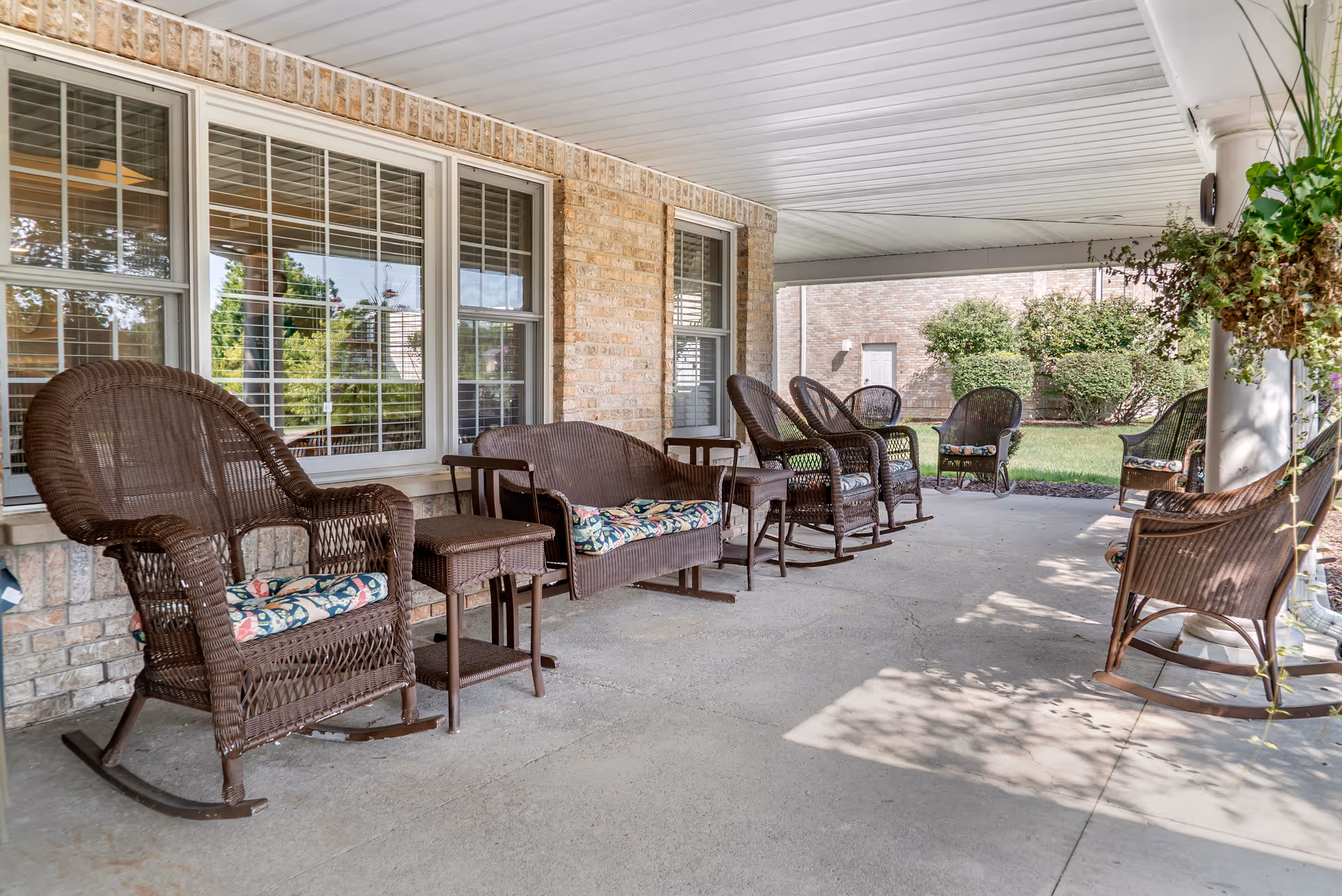 Covered outdoor patio area with several brown wicker rocking chairs and loveseat with floral cushions, small side tables, and hanging plants. The patio is adjacent to a brick building with large windows and overlooks a grassy area with bushes.