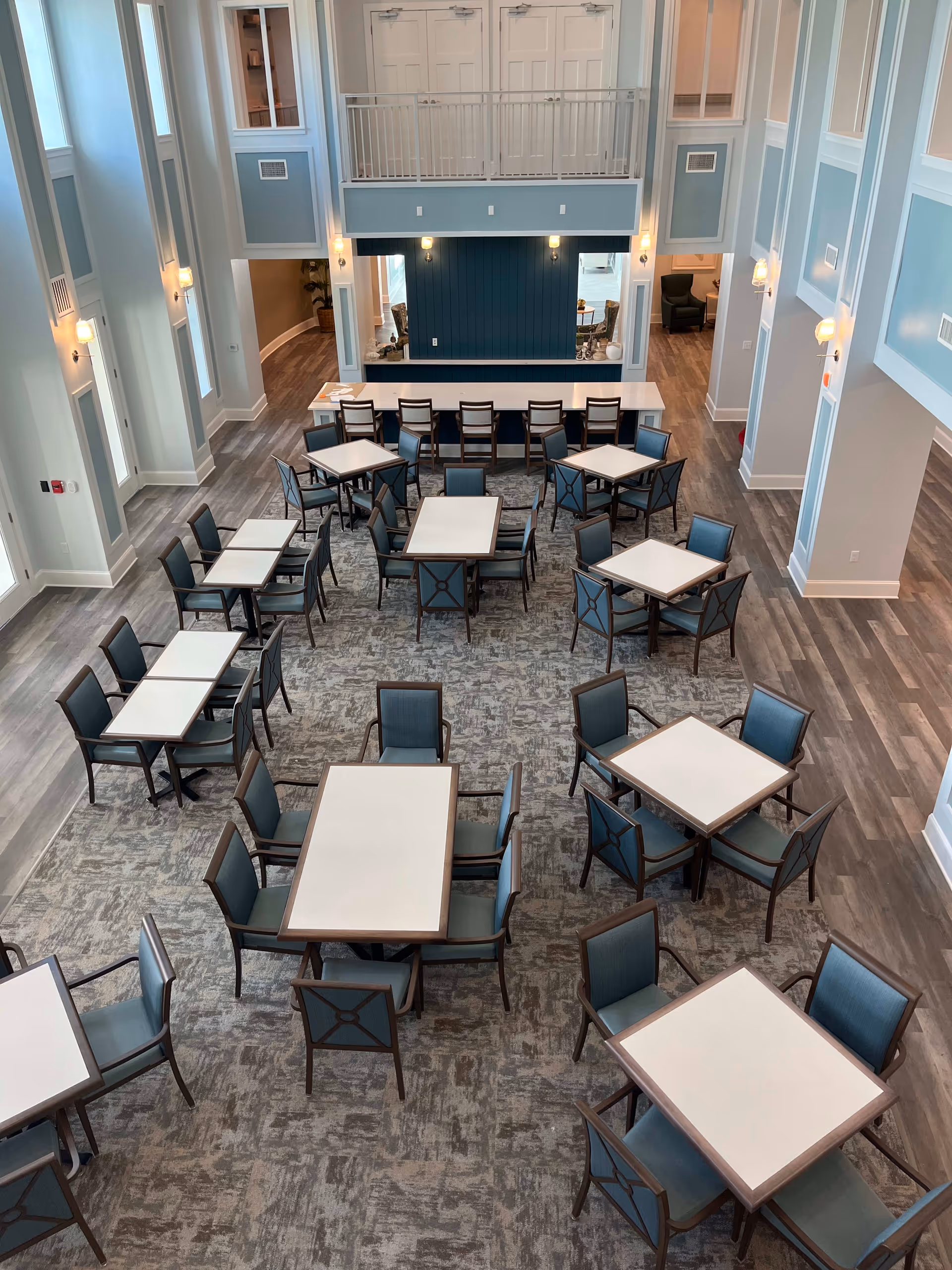 A spacious dining room with multiple square and rectangular tables surrounded by blue cushioned chairs. The room has high ceilings, light blue and white walls, and wood flooring. There is a long counter with bar stools against a dark blue accent wall at the far end of the room. Wall sconces provide lighting along the walls.