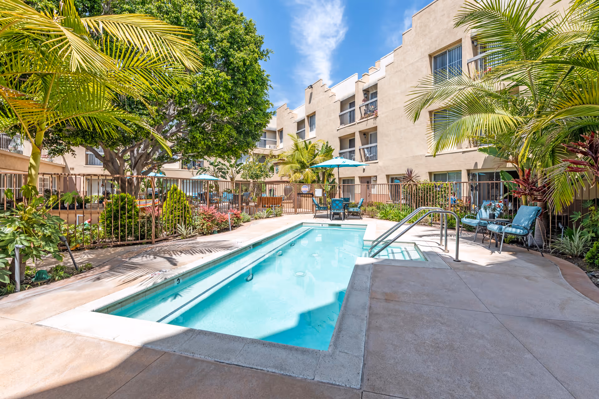 Outdoor swimming pool area surrounded by a beige multi-story building with balconies. The pool is rectangular with metal handrails and steps leading into the water. There are several palm trees and other green plants around the pool area, along with patio chairs and a table with a blue umbrella. The sky is clear with some clouds.