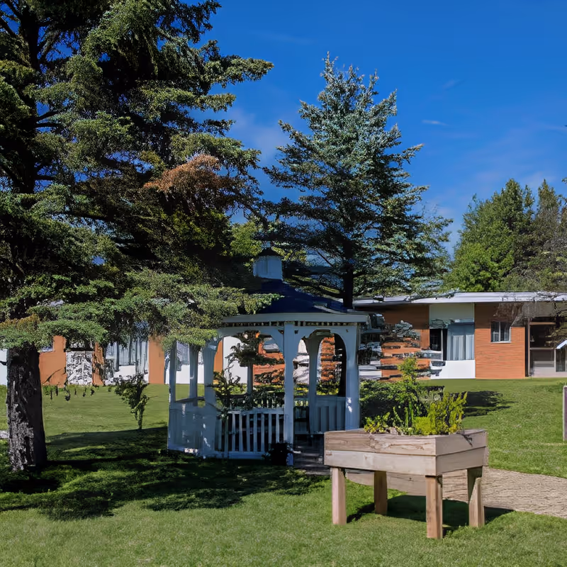 A white wooden gazebo surrounded by green grass and trees in an outdoor garden area of a senior living facility. In the foreground, there is a raised wooden planter box with plants. The building with brick and white walls is visible in the background under a clear blue sky.