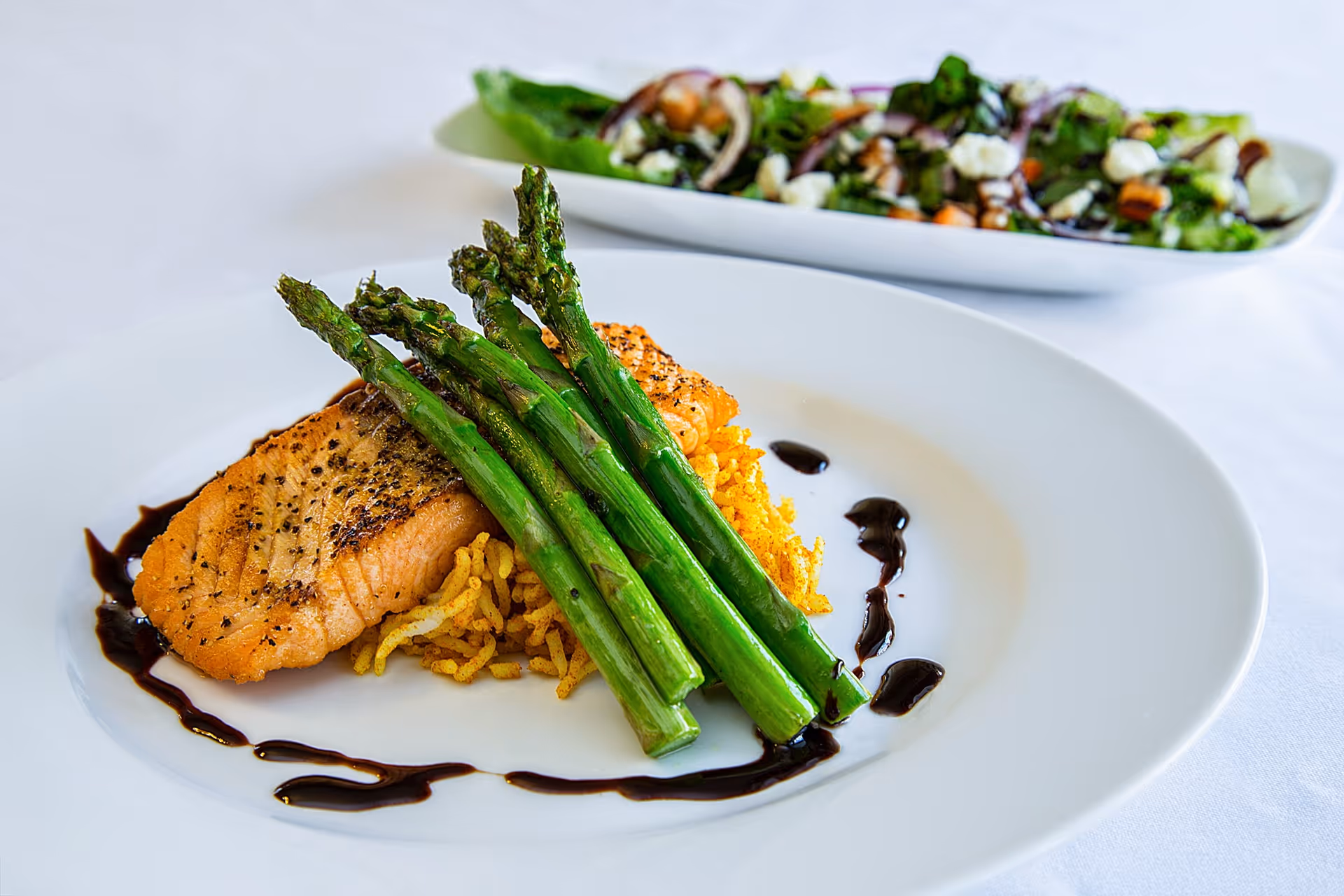 A plated meal featuring grilled salmon topped with several stalks of asparagus, served on a bed of seasoned rice with a drizzle of dark sauce around the edge of the plate. In the background, there is a blurred dish of mixed salad with greens, nuts, and cheese.