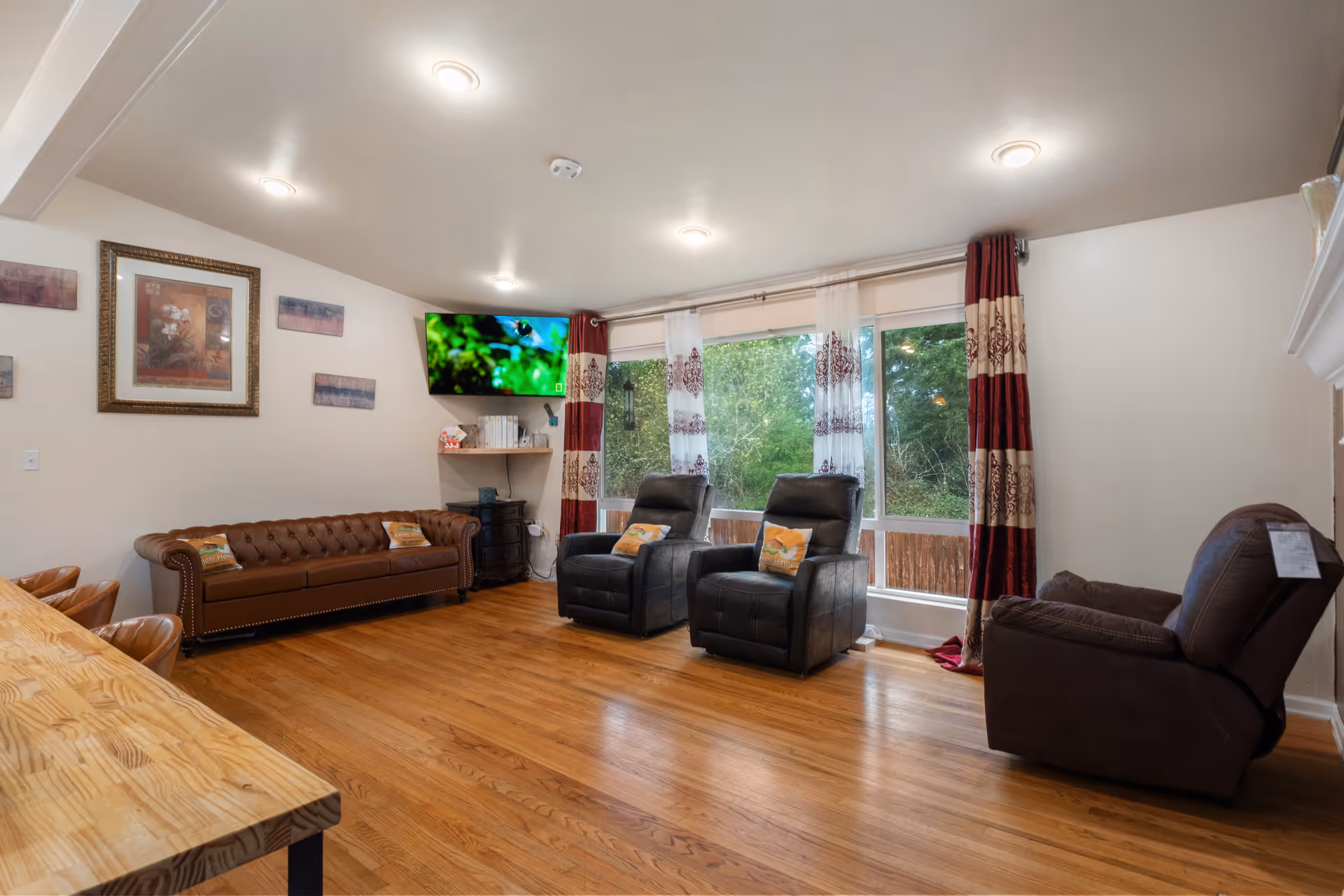 A cozy living room with wooden flooring, featuring a brown leather sofa, two black recliner chairs with decorative pillows, and a brown recliner chair. A flat-screen TV is mounted on the wall above a small shelf with books and decor. Large windows with red and white patterned curtains allow natural light to brighten the room. The walls are decorated with framed artwork.