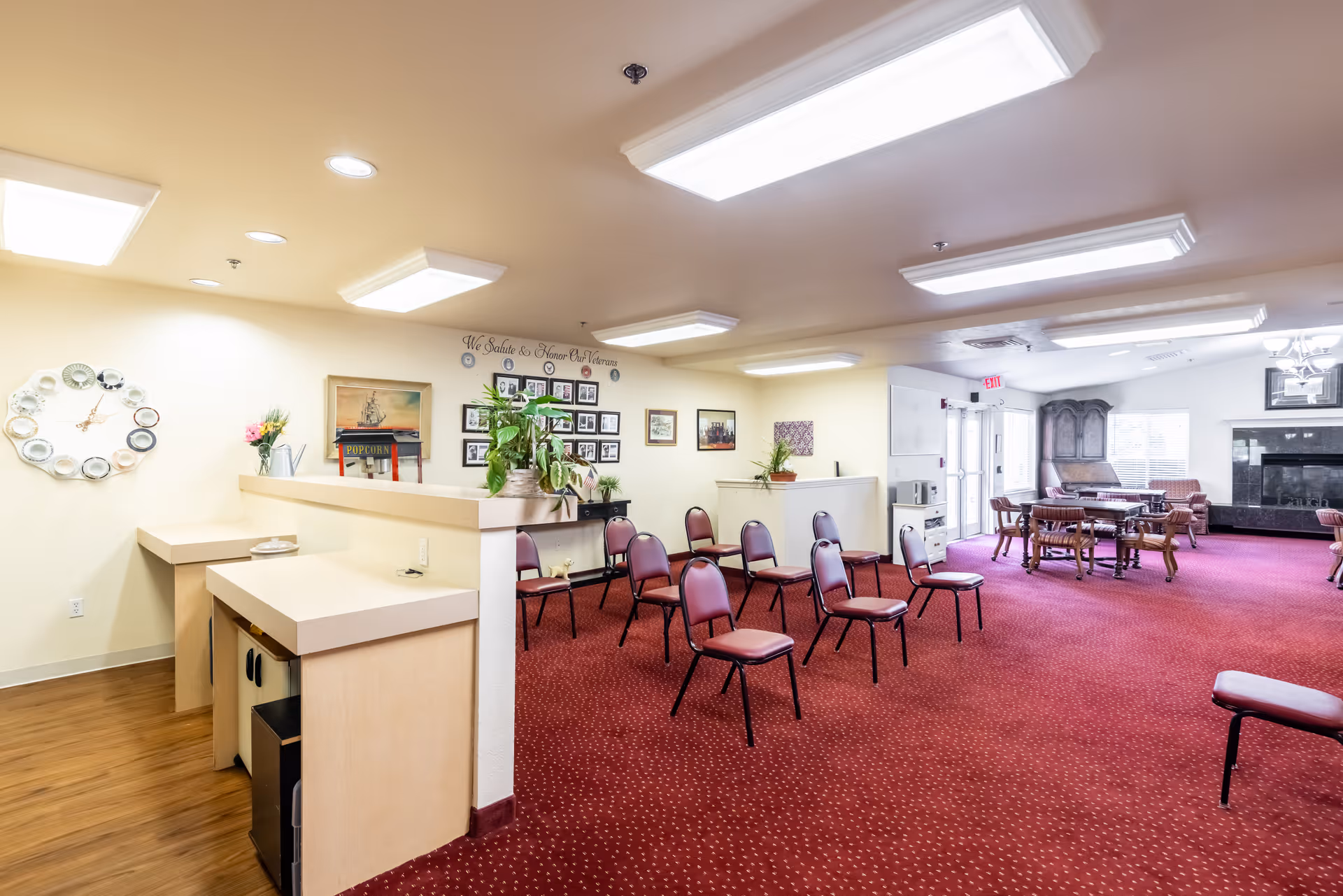 A spacious senior living common area with red carpet and multiple chairs arranged in rows and around tables. The room features a fireplace, a popcorn machine, wall decorations including a clock made of plates, framed photos, and plants. The area is well-lit with ceiling lights and has an exit door with windows.