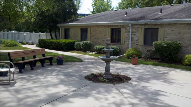 Outdoor courtyard area with a concrete walkway, a three-tiered water fountain in the center, a wooden bench with a memorial inscription, potted plants, and a brick building with windows and shutters in the background.