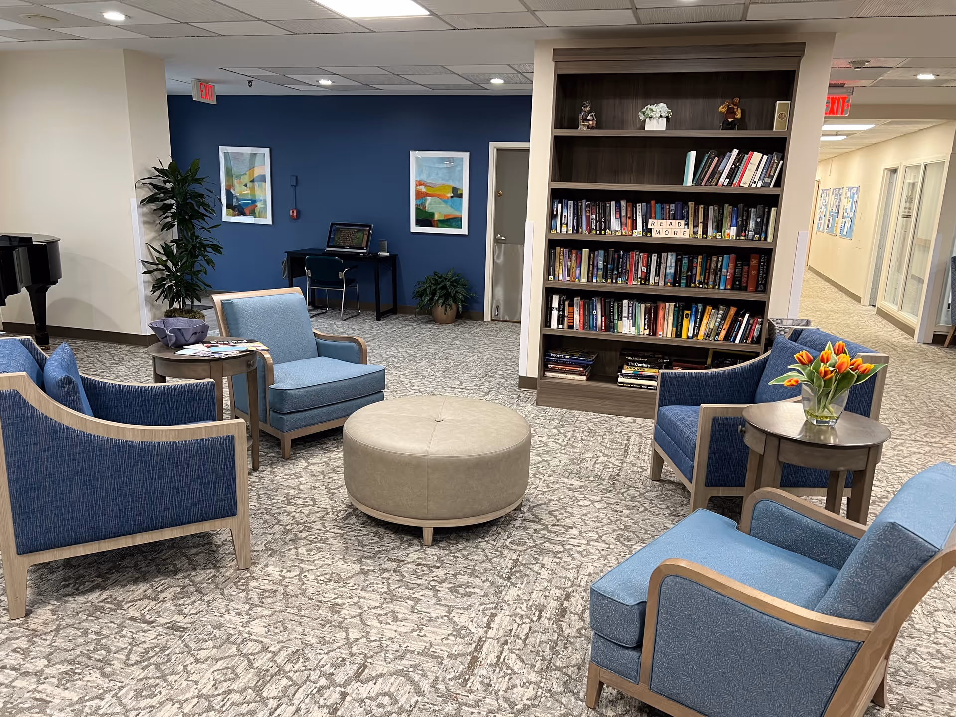 A cozy common area in Myers Apartments Independent Living featuring blue upholstered chairs arranged around a round beige ottoman. There is a wooden bookshelf filled with books and a sign that says 'READ MORE'. A small table with a vase of orange tulips is beside one of the chairs. In the background, there is a desk with a computer, two framed abstract paintings on a blue accent wall, and some potted plants. The area is well-lit with ceiling lights and has a patterned carpet floor.