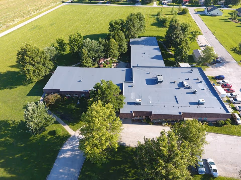Aerial view of a single-story building with a flat roof surrounded by green lawns, trees, and a parking area with several cars. The building is situated in a suburban area with residential houses visible in the background.