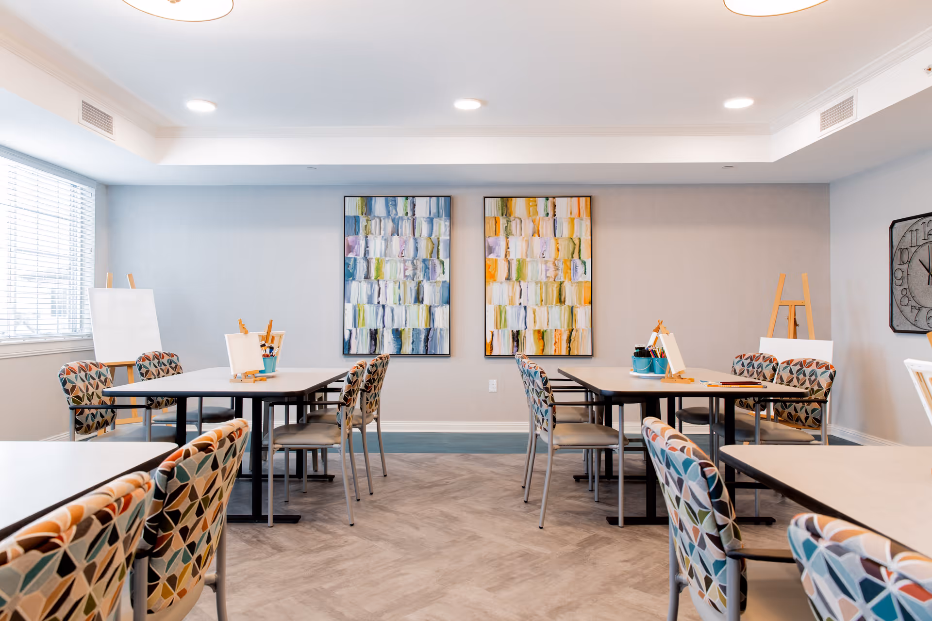 Bright senior living activity room with tables, patterned chairs, easels, and colorful abstract paintings on the wall.