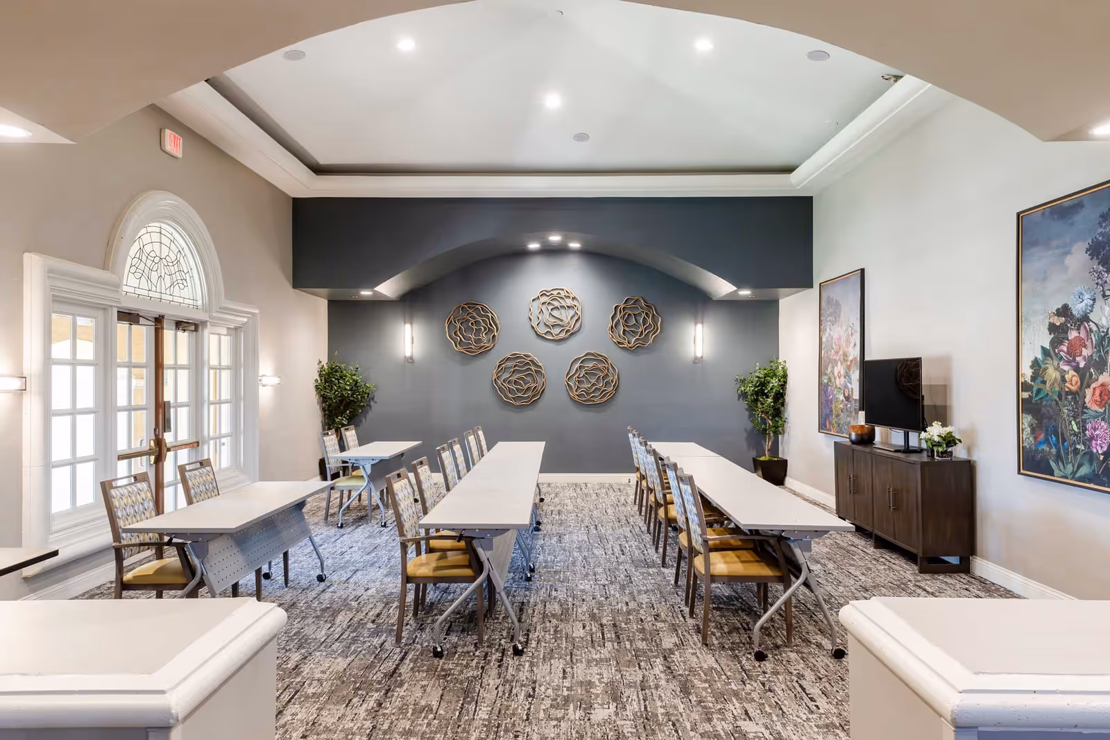 Bright, formal meeting room with long tables and chairs, decorative wall art, a TV cabinet, and large arched windows.