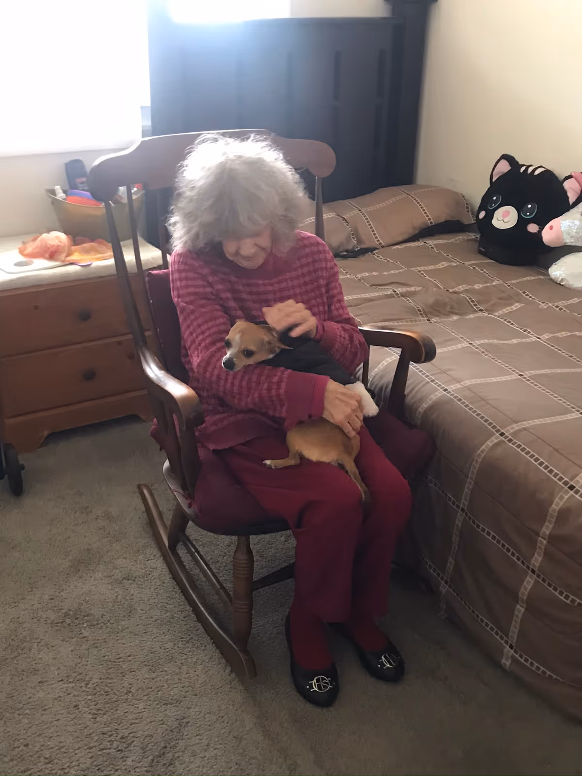 An elderly woman with gray hair wearing a red and pink striped sweater and maroon pants sits in a wooden rocking chair in a bedroom, holding and petting a small brown and white dog. Behind her is a bed with a brown patterned bedspread and two plush toys, one resembling a black cat and the other a white animal. A wooden dresser with items on top is visible to the left.
