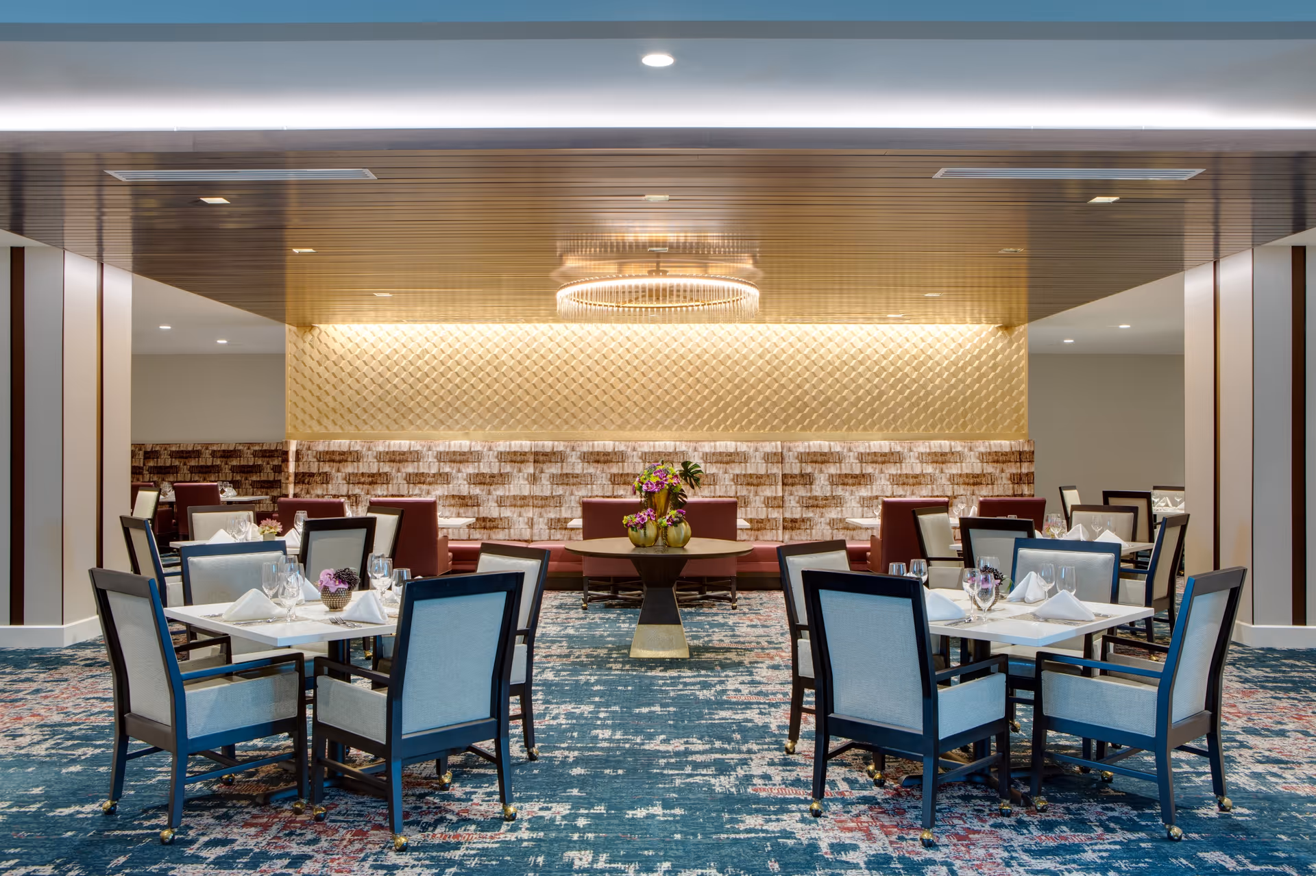 A modern dining room with multiple tables and chairs arranged neatly. Each table is set with white napkins, glassware, and small floral centerpieces. The room features a patterned blue carpet, a textured wall with warm lighting, and a decorative chandelier hanging from a wooden ceiling panel.