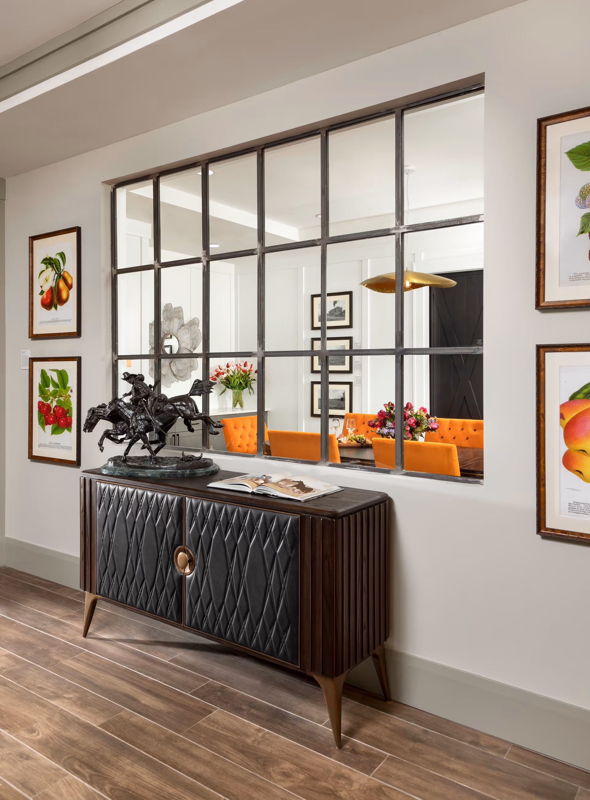 A stylish interior hallway with a dark wood and leather console table topped by a horse sculpture, set before a metal-framed window that looks into a dining area with orange chairs and floral arrangements.