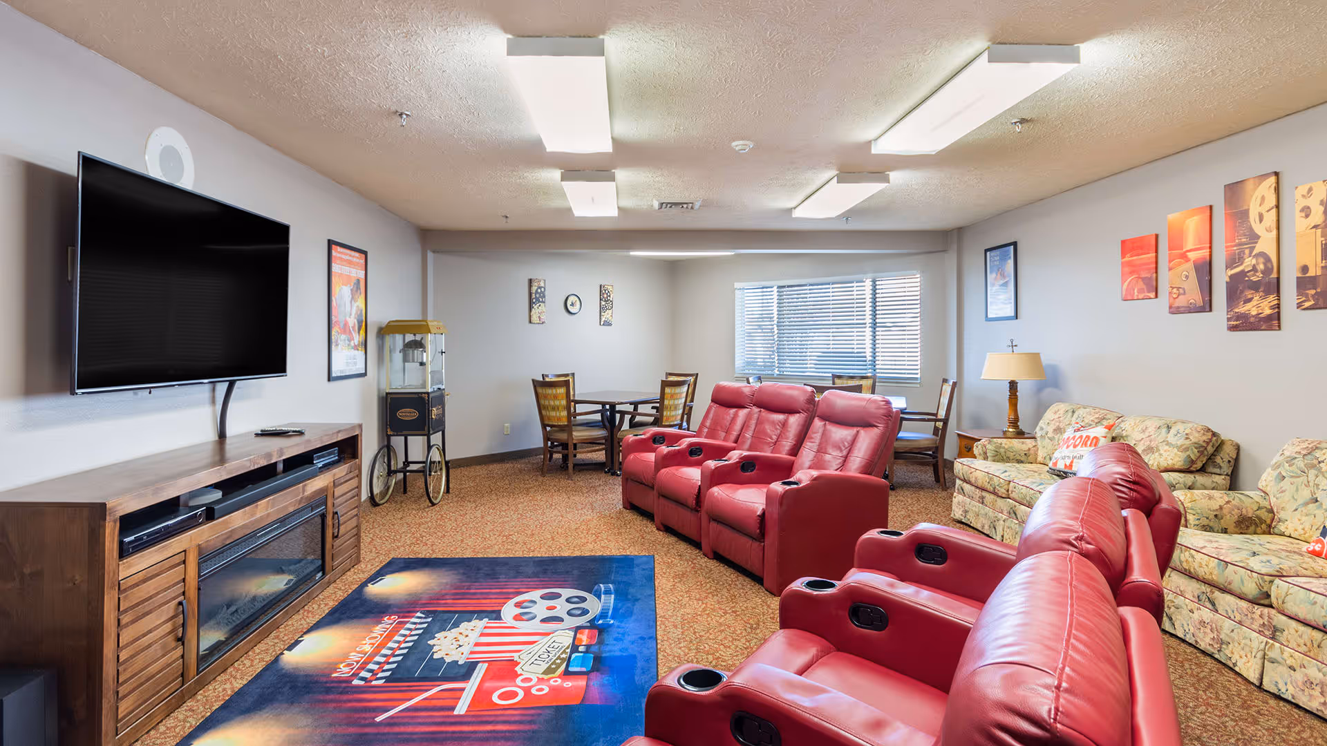 A cozy common area in a senior living facility featuring a large flat-screen TV mounted on the wall above a wooden entertainment center with an electric fireplace. In front of the TV are two rows of red leather recliner chairs with cup holders. To the right, there are floral-patterned sofas and a side table with a lamp. The room has carpeted floors, a popcorn ceiling with fluorescent lights, and several framed movie-themed artworks on the walls. A popcorn machine and a small dining table with chairs are visible in the background near a window with blinds.