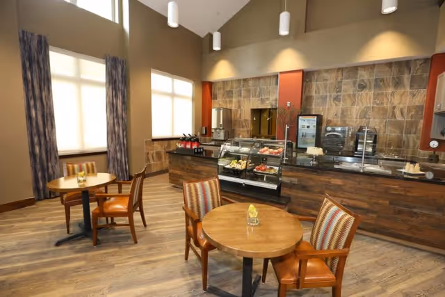 A dining area in a senior living facility with round wooden tables and cushioned chairs. The room has large windows with curtains, wood flooring, and a counter with a glass display case containing food items. The walls behind the counter are decorated with stone tiles and there are pendant lights hanging from the ceiling.