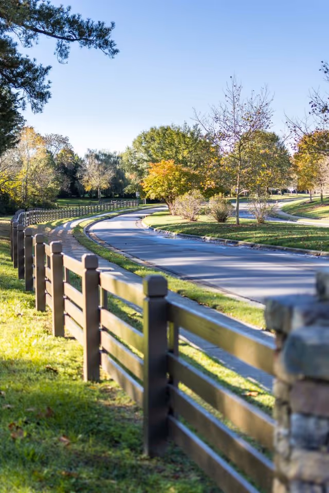 A winding paved road bordered by a wooden fence on one side and grassy areas with trees showing autumn colors on both sides under a clear blue sky.