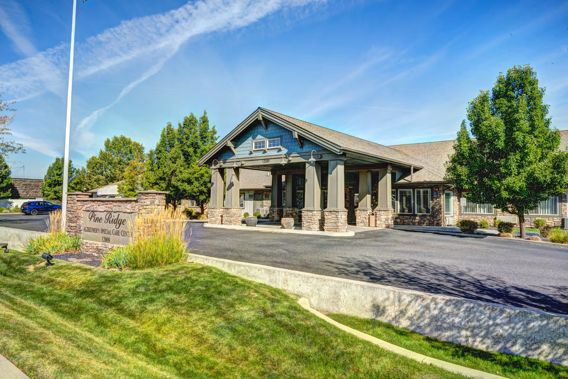 Exterior view of Pine Ridge Alzheimer's Special Care facility showing the entrance with a covered driveway, stone pillars, a flagpole with an American flag, green trees, and a well-maintained lawn under a blue sky.