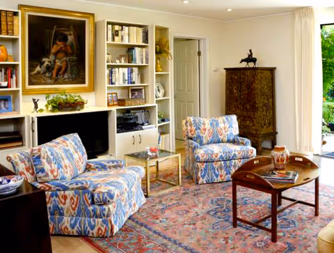 A cozy living room with two patterned armchairs and a matching loveseat arranged around a wooden coffee table on a large decorative rug. The room features built-in white bookshelves filled with books and decorative items, a painting above the fireplace, and a tall ornate cabinet. Large windows with light curtains allow natural light to brighten the space and provide a view of greenery outside.