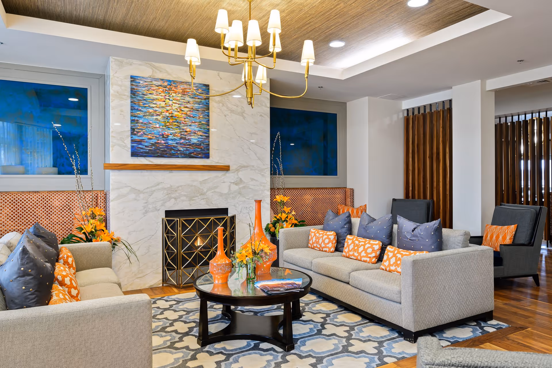 A modern and cozy living room with two beige sofas adorned with orange and blue patterned pillows, a round glass coffee table with orange vases and flowers, a marble fireplace with a decorative screen, and a colorful abstract painting above it. The room features wooden flooring, a patterned area rug, a gold chandelier, and wooden slatted room dividers.