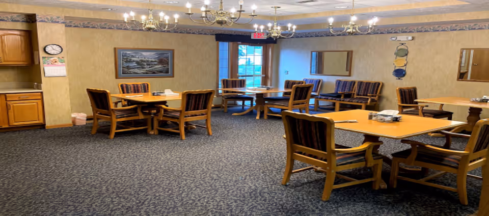 Dining room/common area with multiple wooden tables and upholstered chairs, chandeliers, and a small kitchenette area.