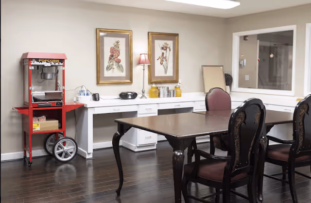 A room with a dark wooden table surrounded by four chairs with dark wood and maroon cushions. Against the wall is a white desk with drawers, decorated with two framed floral paintings, a small lamp, a radio, and some books. To the left, there is a red popcorn machine on wheels. The floor is dark hardwood, and the walls are light-colored.