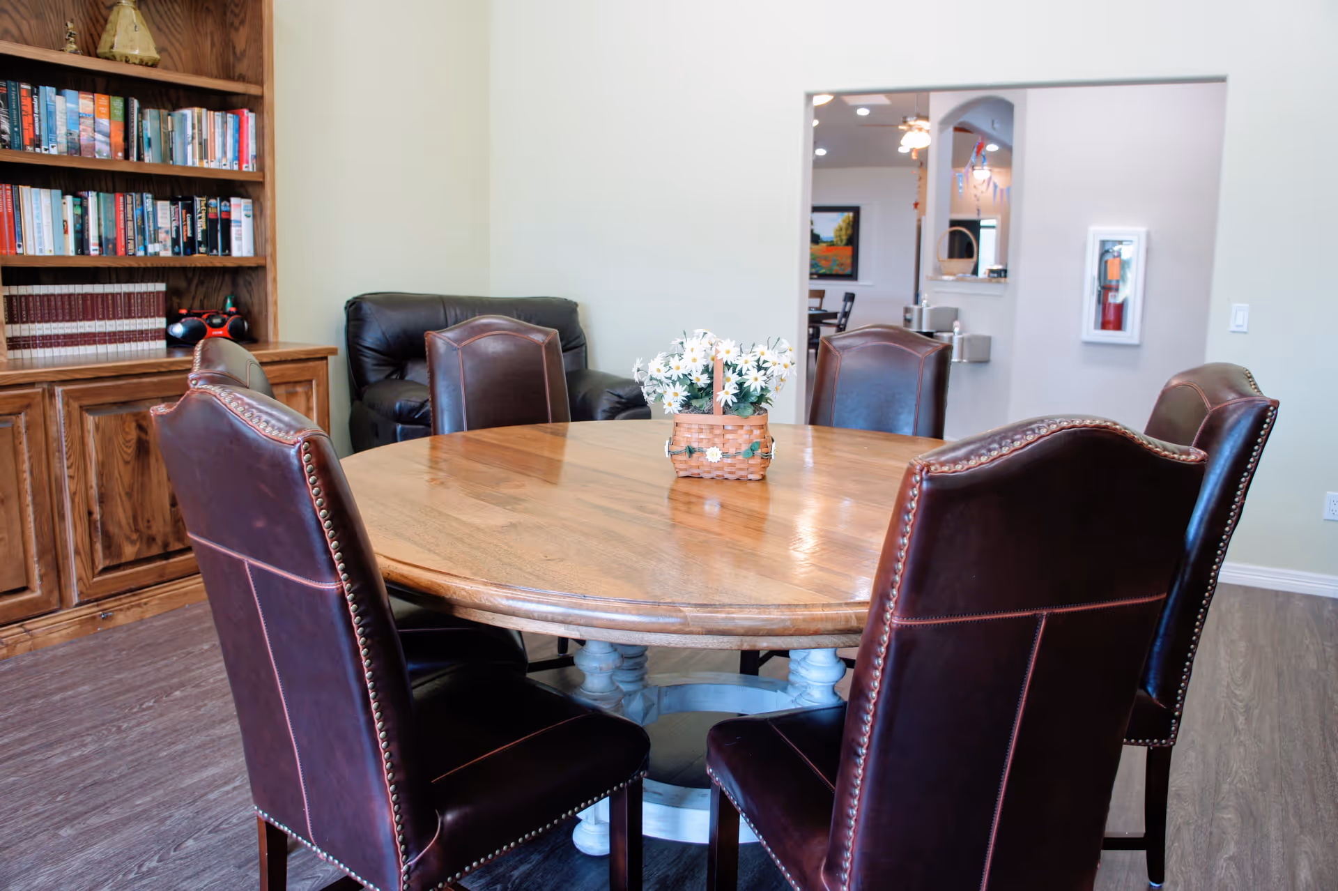 A round wooden dining table with a small basket of white daisies in the center, surrounded by six dark brown leather chairs with nailhead trim. In the background, there is a wooden bookshelf filled with books and a black leather armchair. The room has light-colored walls and wood flooring, with a view into another room through a rectangular opening in the wall.