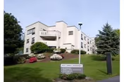 Exterior view of a multi-story senior living facility building named Marywood Heights, surrounded by landscaped greenery including bushes, trees, and a lawn. A sign with the facility name is visible near the entrance driveway.
