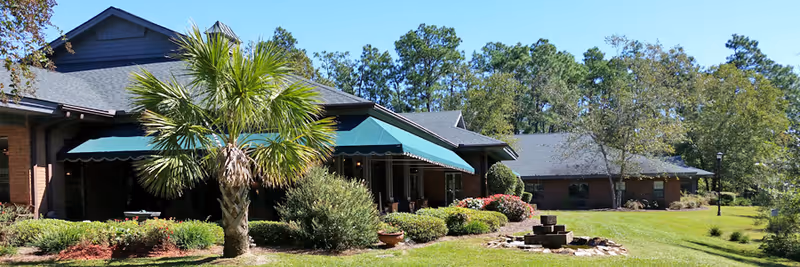 Exterior view of a single-story brick assisted living facility with green awnings over the windows, surrounded by well-maintained landscaping including a palm tree, bushes, and a grassy lawn with trees in the background under a clear blue sky.