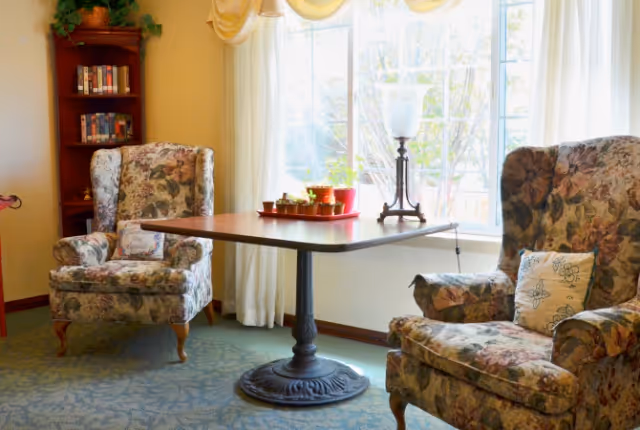 A cozy sitting area with two floral upholstered armchairs facing a wooden table with a decorative lamp and small potted plants on it, next to a large window with sheer curtains letting in natural light. A small bookshelf with books is visible in the corner.
