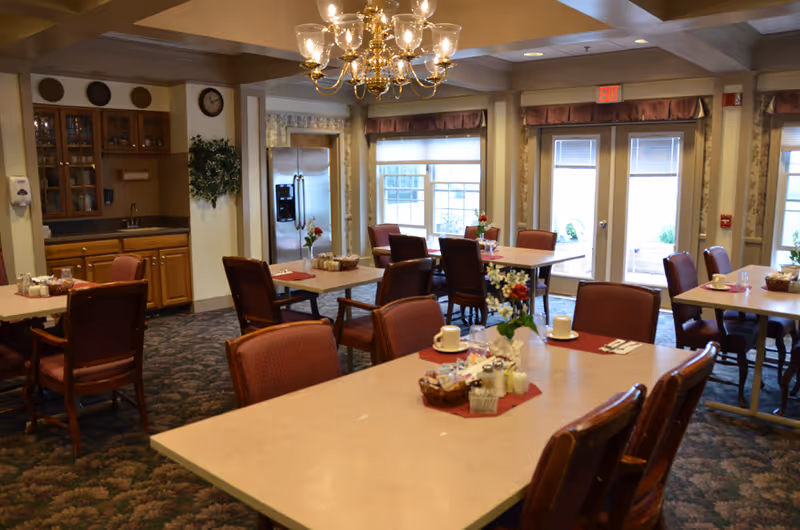 A dining room in a retirement community with several tables and chairs arranged neatly. Each table has a centerpiece with flowers, cups, and condiments. The room features a chandelier, large windows with curtains, and a stainless steel refrigerator near a small kitchenette area with wooden cabinets and a sink.