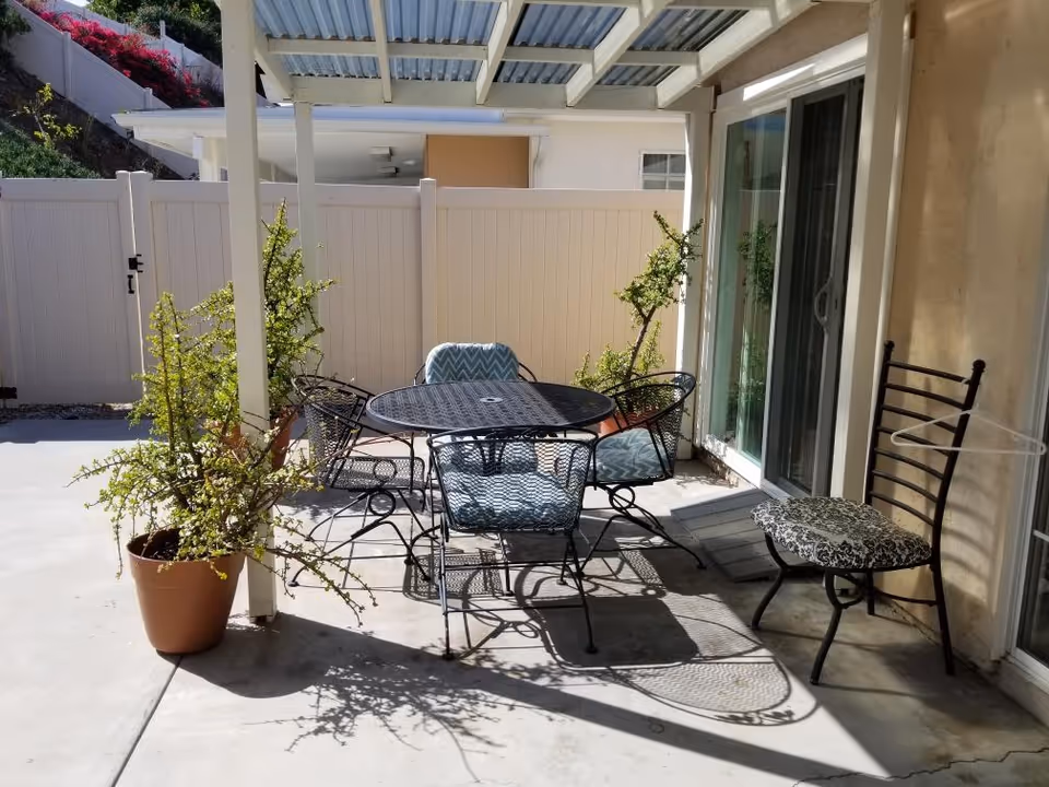 Covered patio with a round metal table, several metal chairs with cushions, potted plants, and sliding glass doors.