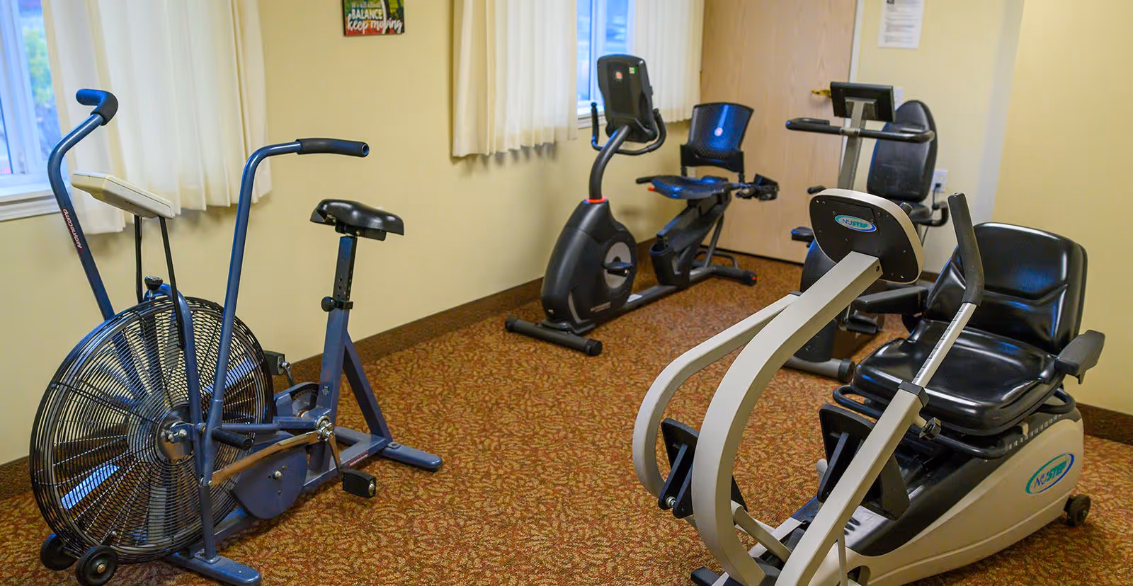 A small exercise room with three stationary exercise bikes on a patterned carpet floor. The room has beige walls, a window with cream curtains, and a motivational poster on the wall.