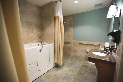 A spacious bathroom with a walk-in bathtub on the left side, beige tiled walls and floor, a shower curtain, a green painted wall, a granite countertop with a sink, a mounted soap dispenser, a towel rack with white towels, and a toilet paper holder.