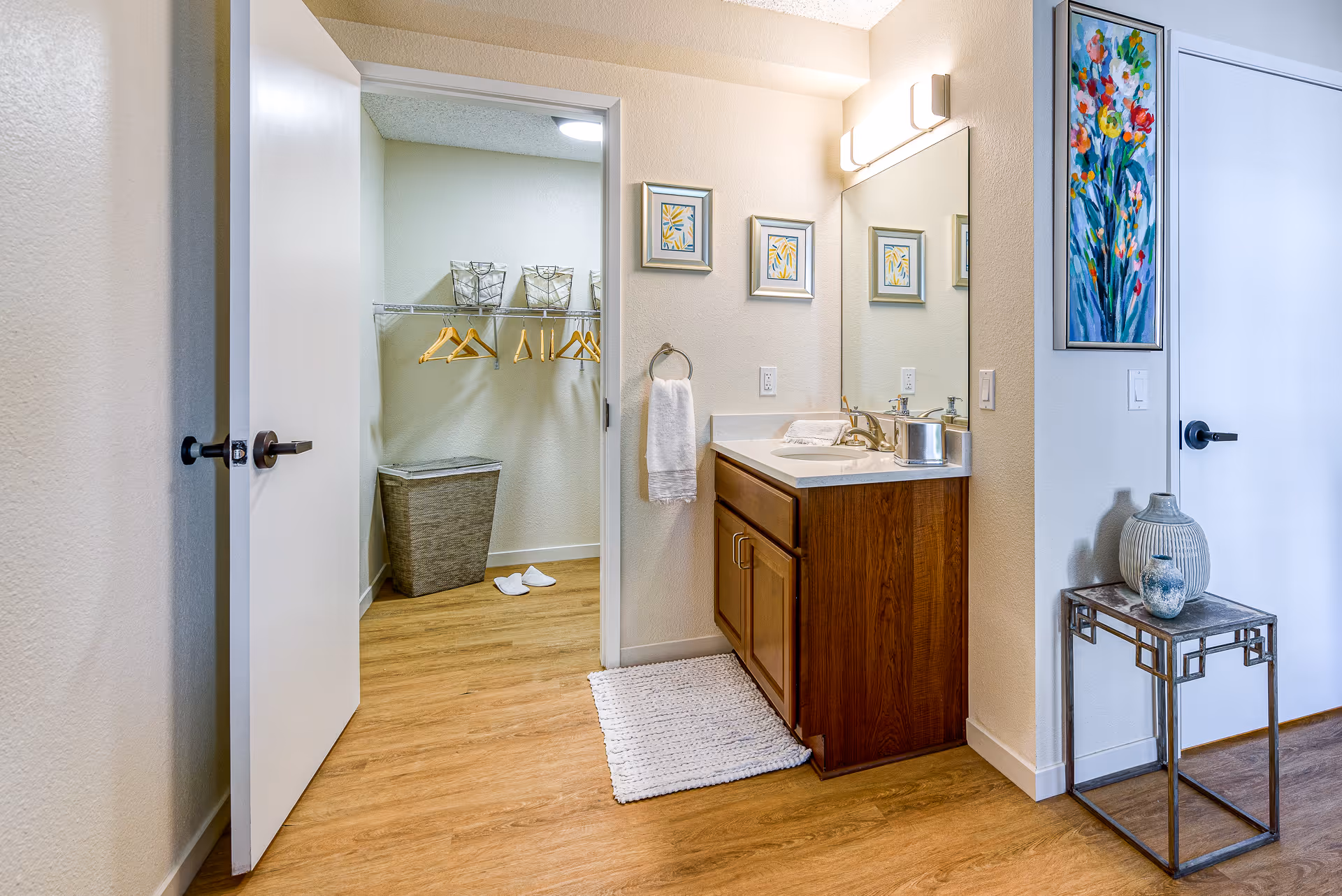Vanity with sink and mirror beside an open walk-in closet with hangers and a decorative side table in a senior living apartment.