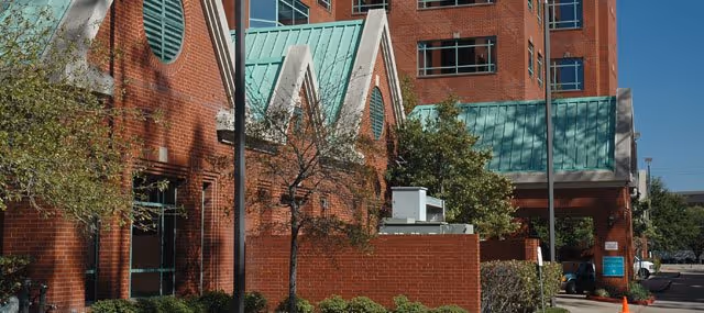 Exterior view of a red brick building with green metal roofs and multiple windows, surrounded by trees and shrubs under a clear blue sky.