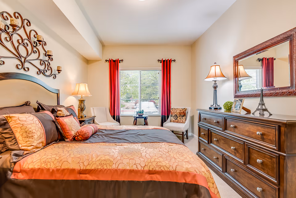 Furnished bedroom with a large bed, decorative wall art, dresser with mirror, lamps, and a window with red curtains.