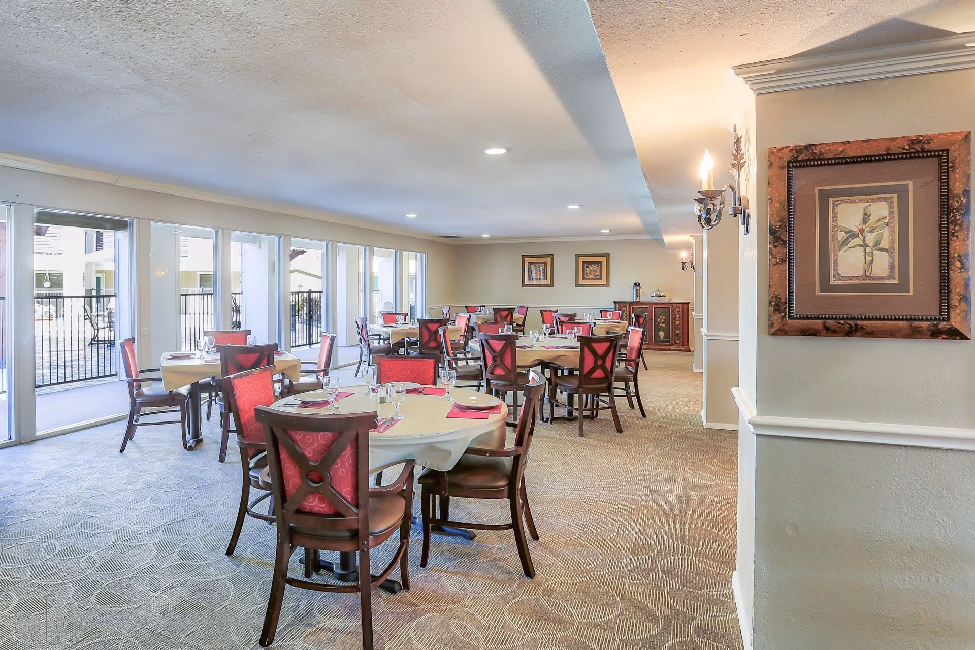 Dining room with round tables set for a meal, red-upholstered chairs, and large windows along one wall.