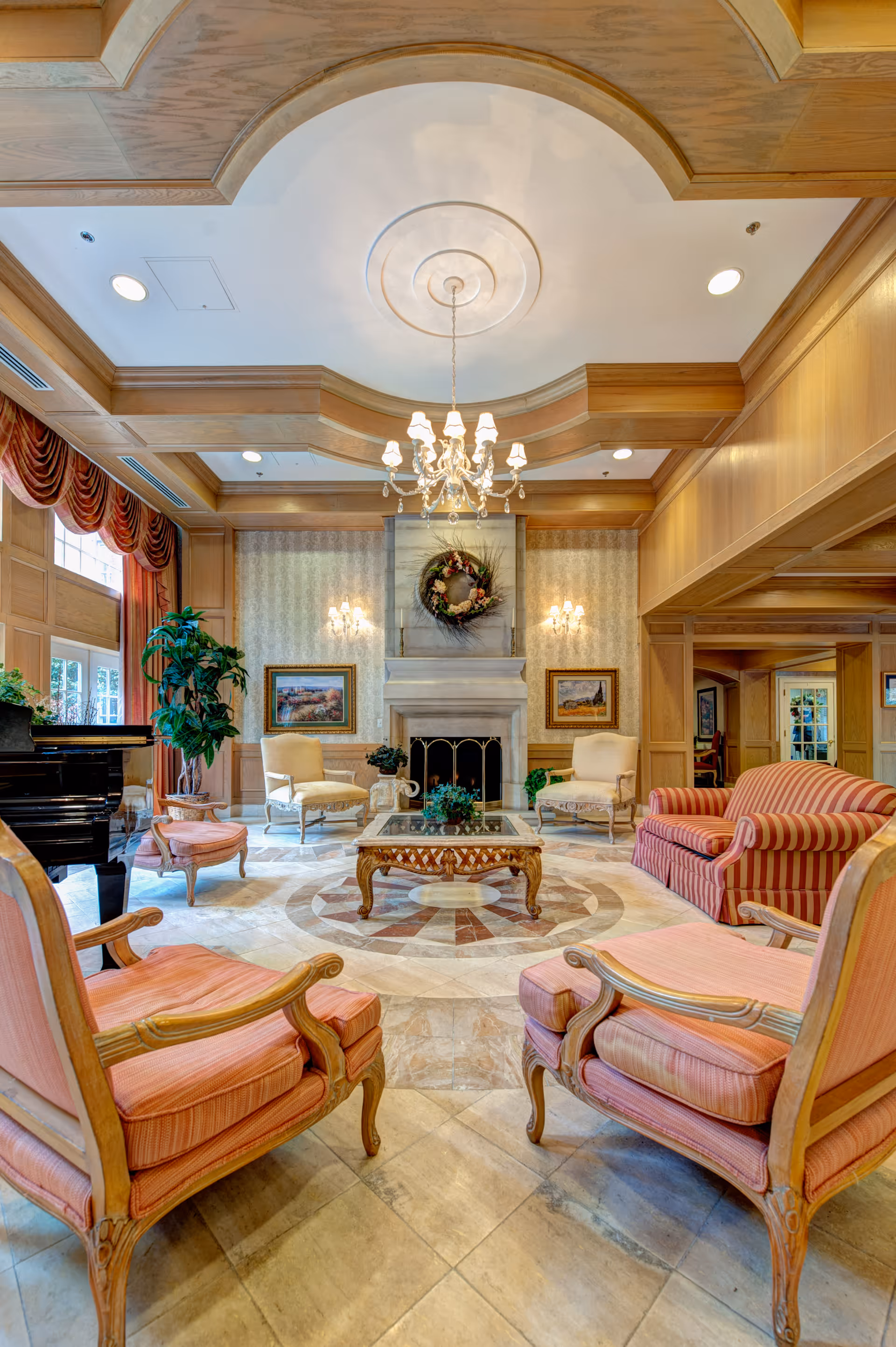 A spacious and elegant living room with a high ceiling featuring a chandelier. The room has a fireplace adorned with a wreath, flanked by two wall sconces and framed paintings. There are multiple upholstered chairs and a striped sofa arranged around a glass coffee table on a decorative tiled floor. Large windows with red drapes allow natural light to fill the room, and a black grand piano is visible on the left side.