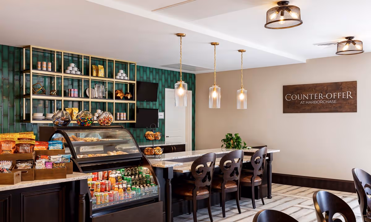 Interior view of a snack and beverage counter area in a senior living facility. The counter has a glass display case filled with pastries and refrigerated drinks below. Shelves behind the counter hold various snacks, cups, and decorative items. There are three pendant lights hanging above a marble countertop with four wooden chairs. A wooden sign on the beige wall reads 'COUNTER-OFFER AT HARBORCHASE'.
