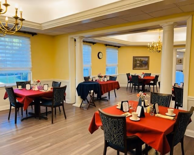 A dining room in a senior living facility with several tables covered in red and dark blue tablecloths. Each table is set with cups, plates, utensils, and small flower arrangements. The room has yellow walls, large windows with white blinds, wooden flooring, and chandeliers hanging from the ceiling. White columns separate different sections of the room.