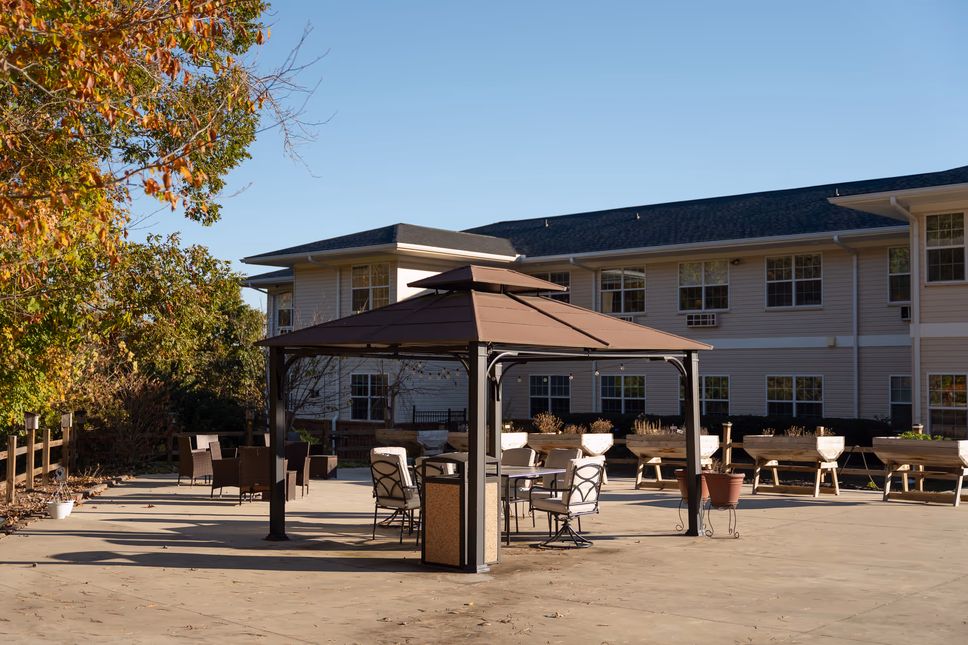Outdoor patio area at Oaks at Ellijay featuring a brown gazebo with cushioned chairs and a table underneath. Surrounding the patio are wooden planter boxes and additional seating. The background shows a two-story building with multiple windows and a clear blue sky.