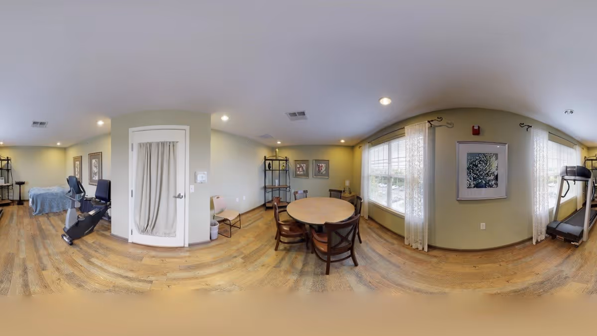 A spacious, well-lit room with light green walls and wooden flooring. The room features a round wooden table with four chairs near large windows with sheer white curtains. On one side, there is a treadmill and on the other side, an exercise bike next to a bed covered with a blue blanket. The room also has framed artwork on the walls and a white door with a curtain. Ceiling lights illuminate the space.