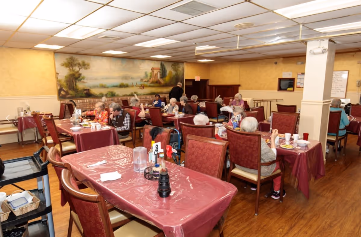 A dining room in a senior living facility with elderly residents seated at tables covered with red tablecloths. The room has wooden floors, beige walls, and a large mural depicting a scenic landscape. Several tables have people eating and socializing.