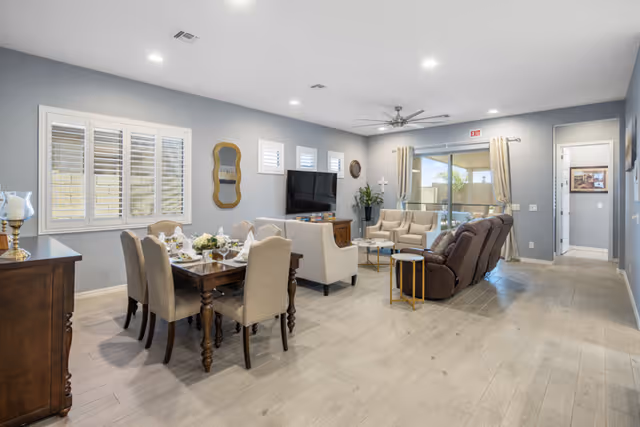 A spacious and well-lit living and dining area in an assisted living facility. The dining area features a wooden table set with six beige upholstered chairs and tableware. The living area has a large flat-screen TV mounted on the wall, two beige armchairs, a brown reclining sofa, and a small round side table. The room has light wood flooring, gray walls, recessed ceiling lights, and large windows with white shutters and beige curtains. A sliding glass door leads to an outdoor patio area.