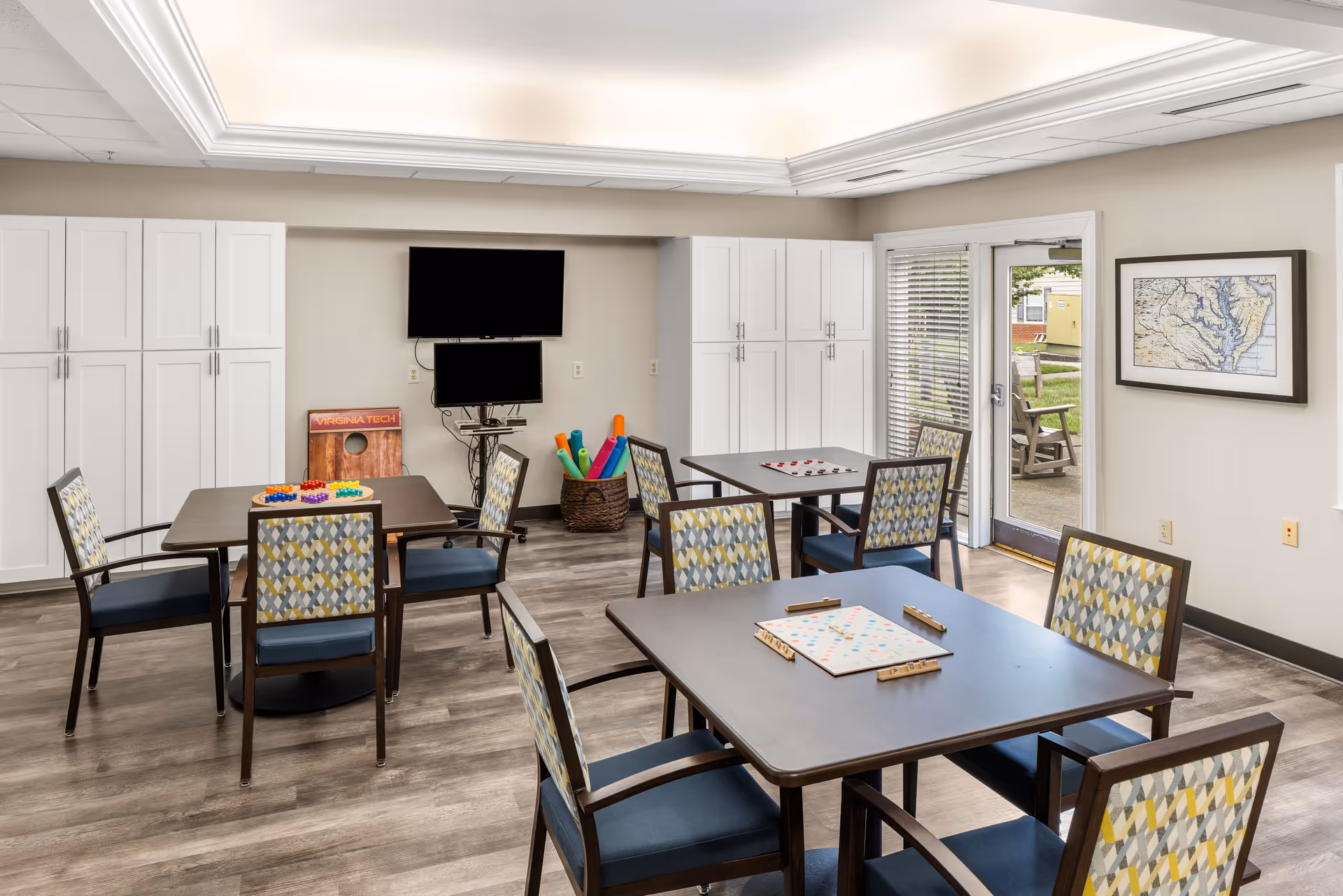 A bright activity room with several tables and chairs arranged for games. One table has a Scrabble board, another has a checkers board, and a third has a colorful peg game. The room features white cabinets along the back wall, two mounted flat-screen TVs, a basket filled with colorful pool noodles, and a door leading outside. A framed map hangs on the wall near the door.